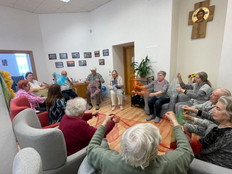 Senioru grupa gaišā telpā sēž aplī un kopīgā nodarbībā rokās tur krāsainus, caurspīdīgus audumus. Pie sienas redzamas ierāmētas fotogrāfijas un reliģiska ikona.