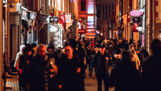 Cilvēku pilna vecpilsētas gājēju iela vakara stundās, ko izgaismo ielu laternas un ēku skatlogi; pie namiem izvietoti Latvijas karogi.