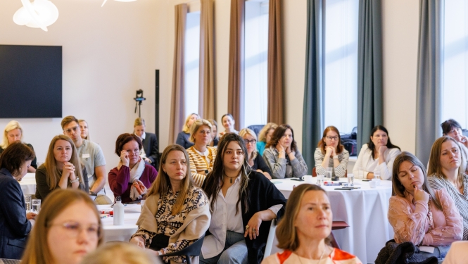 Pasākuma dalībnieki sēž pie galdiem gaišā konferenču telpā un klausās prezentāciju. Zālē redzami vairāki desmiti cilvēku, daļai priekšā ir piezīmju materiāli, datori un dzērieni.