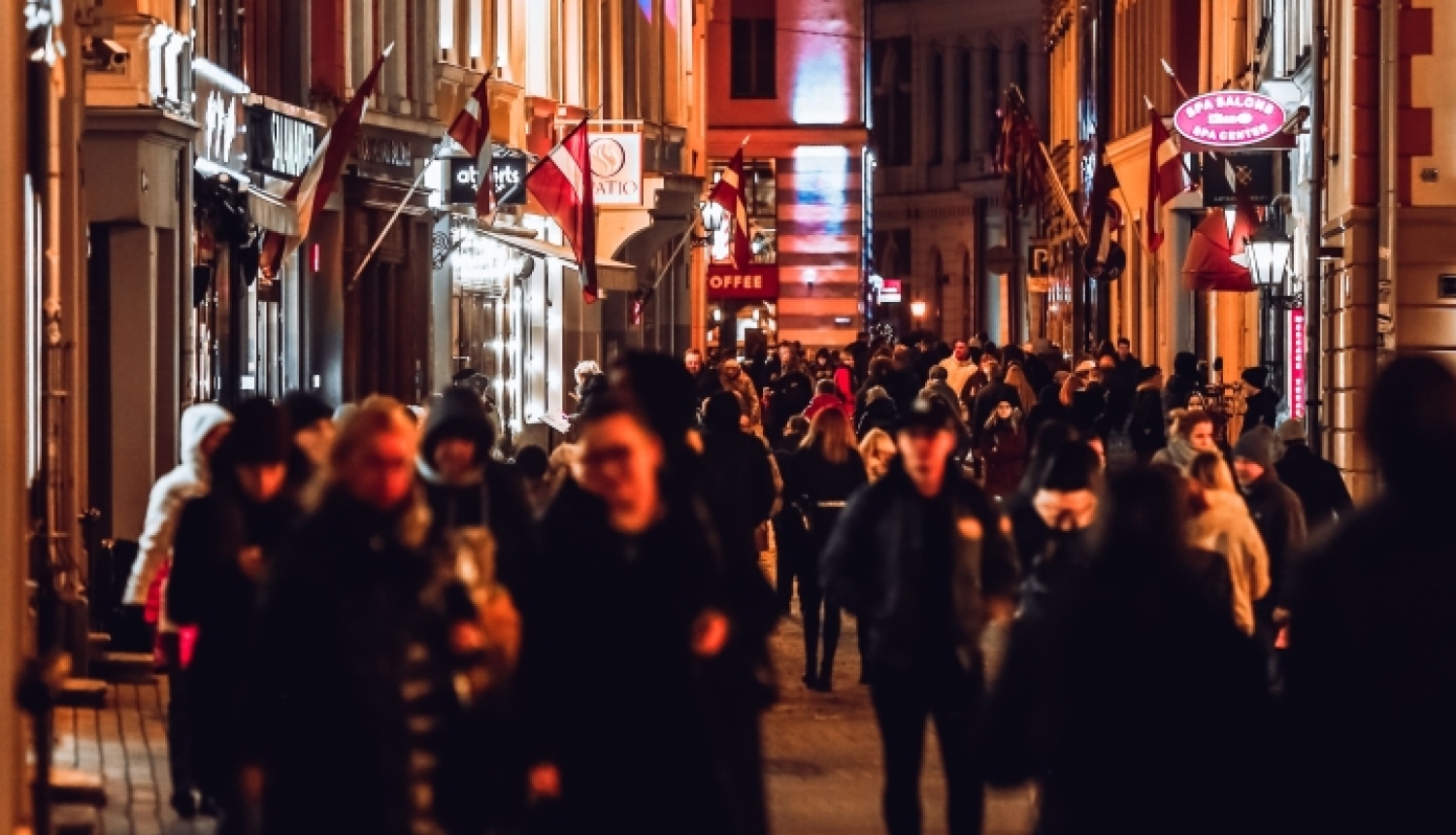 Cilvēku pilna vecpilsētas gājēju iela vakara stundās, ko izgaismo ielu laternas un ēku skatlogi; pie namiem izvietoti Latvijas karogi.