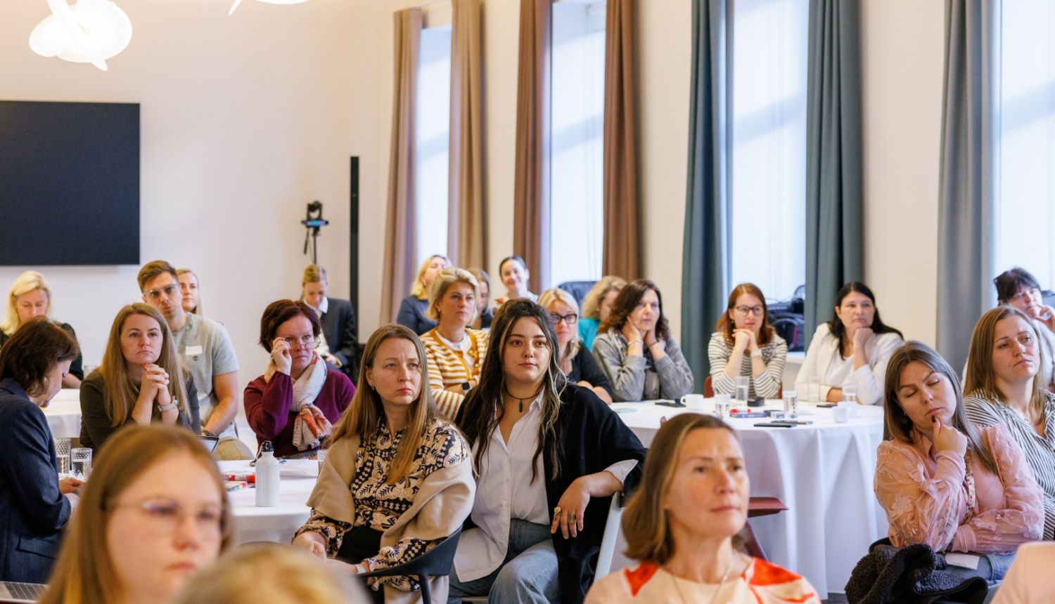 Pasākuma dalībnieki sēž pie galdiem gaišā konferenču telpā un klausās prezentāciju. Zālē redzami vairāki desmiti cilvēku, daļai priekšā ir piezīmju materiāli, datori un dzērieni.