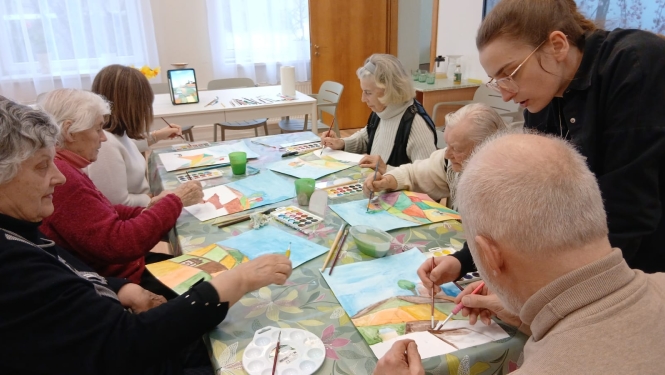 Seniori pie galda piedalās radošajā nodarbībā un ar akvareļkrāsām glezno ainavas. Nodarbības vadītāja palīdz dalībniekiem un sniedz norādes.