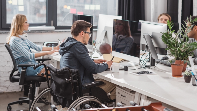 ilustratīva bilde ar cilvēku ar invalidināti ofisā pie galda strādājot blakus kolēģiem.