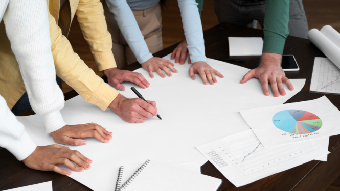 Cilvēku rokas noliktas uz galda virs baltām lapām. Tiek plānots saturs