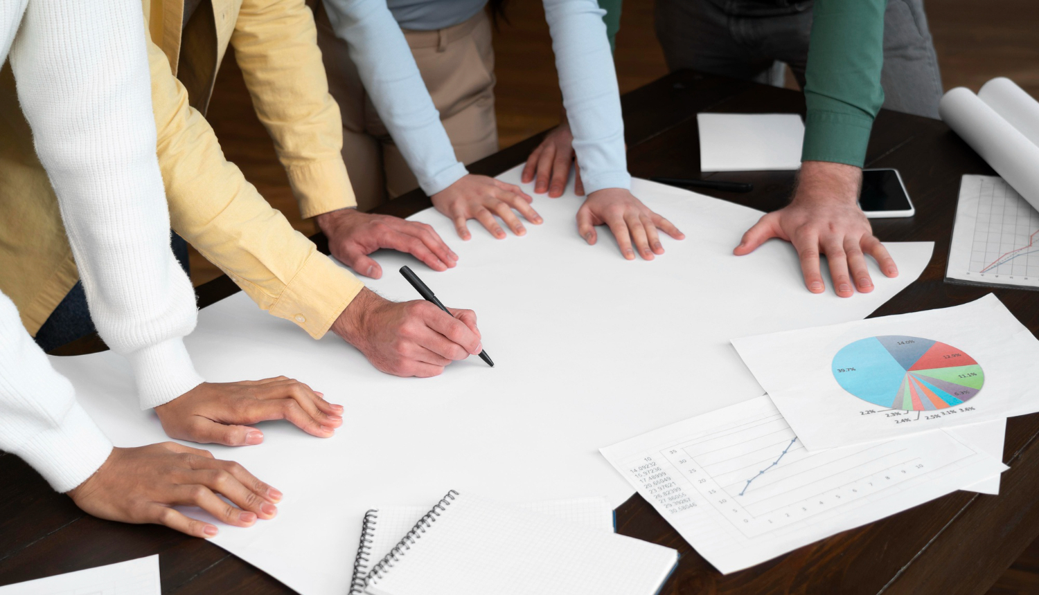 Cilvēku rokas noliktas uz galda virs baltām lapām. Tiek plānots saturs