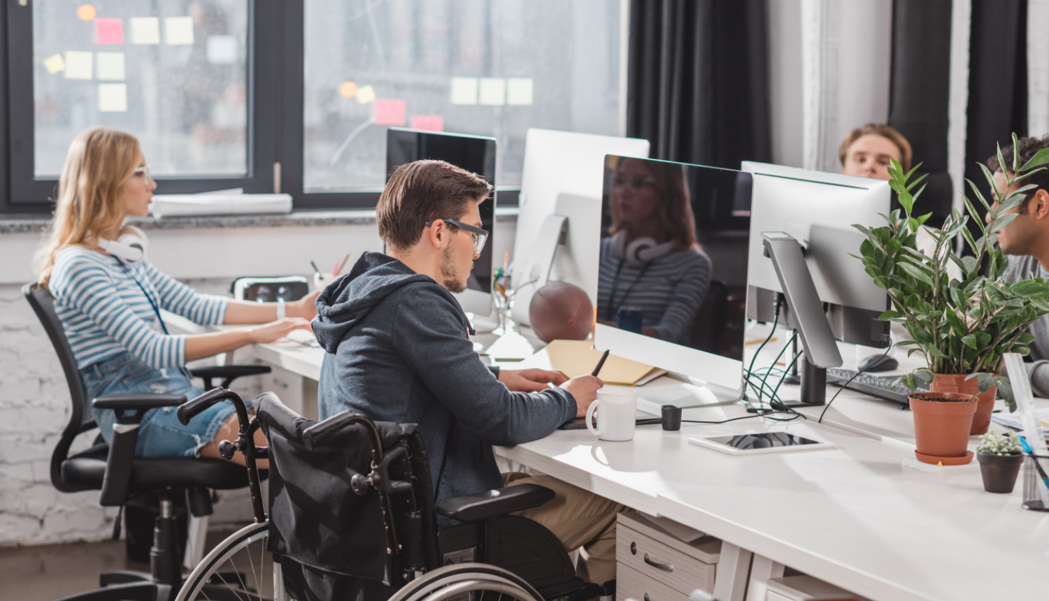 ilustratīva bilde ar cilvēku ar invalidināti ofisā pie galda strādājot blakus kolēģiem.