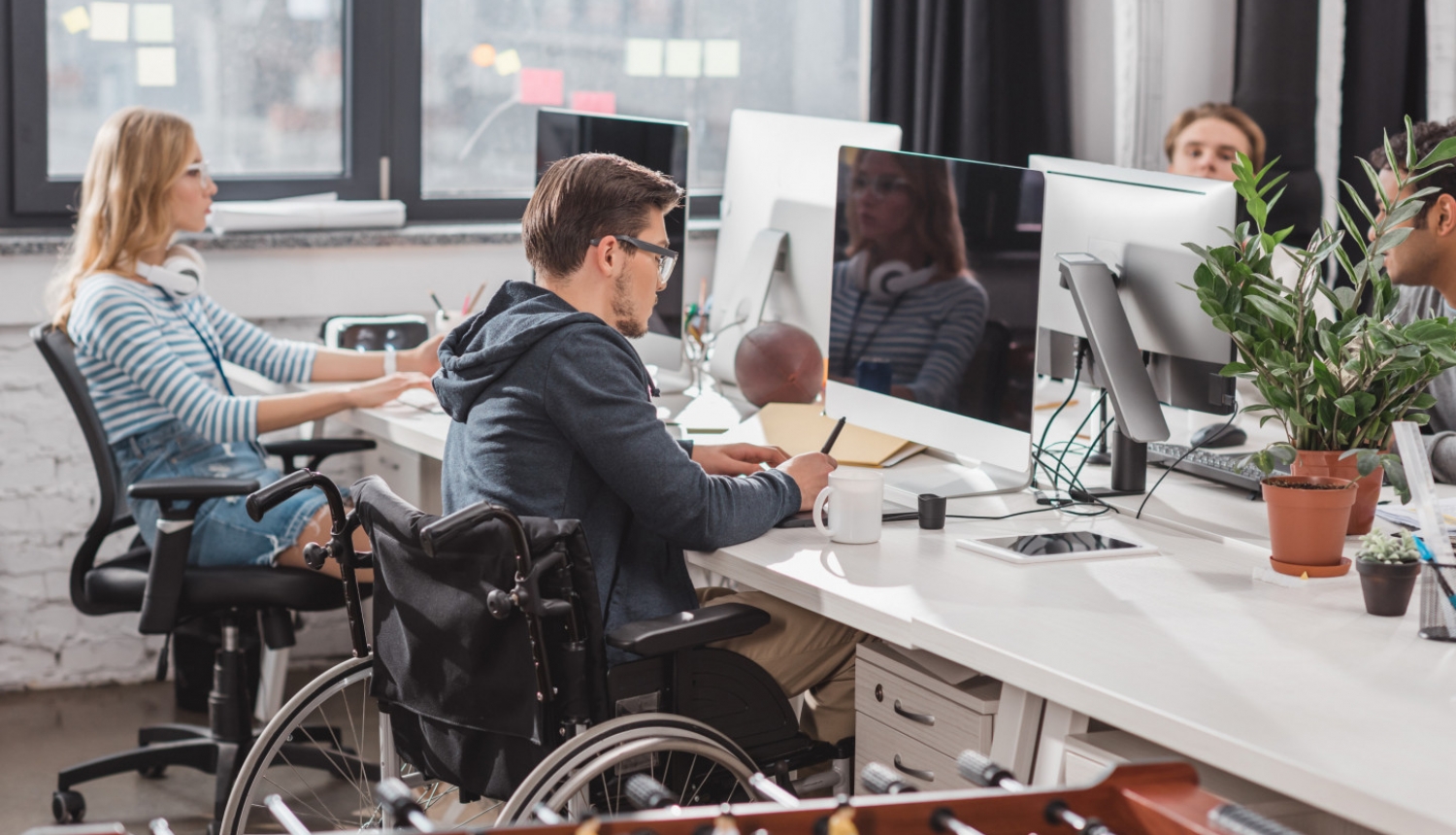 ilustratīva bilde ar cilvēku ar invalidināti ofisā pie galda strādājot blakus kolēģiem.